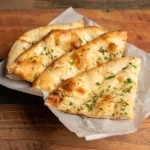 Auf einem Holztisch steht ein Teller, auf dem vier Stücke Naan-Brot aufgereiht sind. Das Brot ist mit kleingehackter Petersilie bestreut.