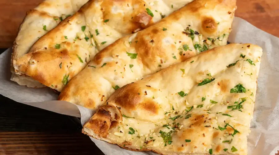 Auf einem Holztisch steht ein Teller, auf dem vier Stücke Naan-Brot aufgereiht sind. Das Brot ist mit kleingehackter Petersilie bestreut.