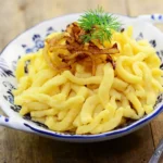 Spätzle in einer weißen Porzellanschüssel mit blauer Bemalung angerichtet mit einer Garnitur aus geschmälzten Zwiebeln und einem Blatt Dill.