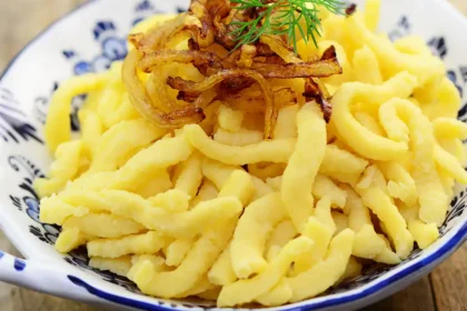 Spätzle in einer weißen Porzellanschüssel mit blauer Bemalung angerichtet mit einer Garnitur aus geschmälzten Zwiebeln und einem Blatt Dill.
