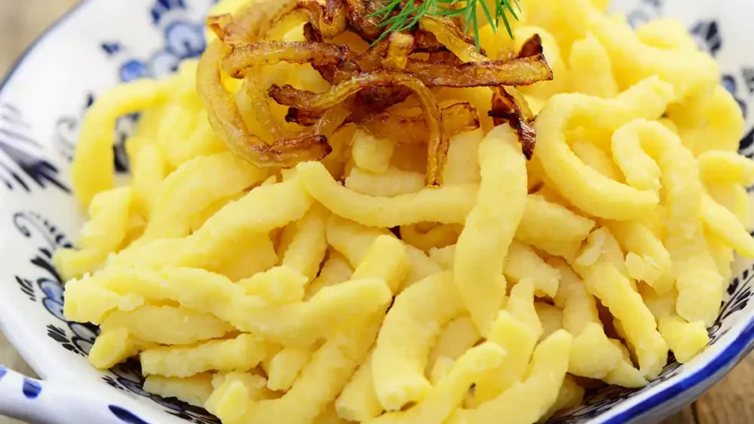Spätzle in einer weißen Porzellanschüssel mit blauer Bemalung angerichtet mit einer Garnitur aus geschmälzten Zwiebeln und einem Blatt Dill.
