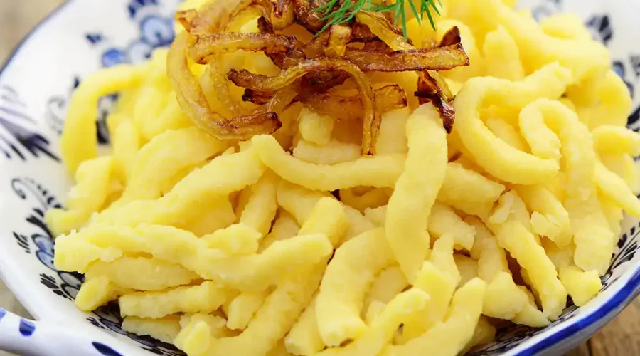 Spätzle in einer weißen Porzellanschüssel mit blauer Bemalung angerichtet mit einer Garnitur aus geschmälzten Zwiebeln und einem Blatt Dill.