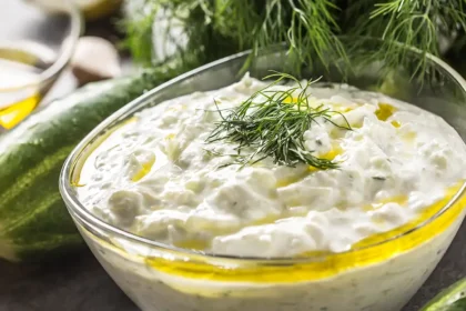 Glasschüssel gefüllt mit Tsatsiki, auf dem Olivenöl schwimmt, garniert mit geschnittenem Dill, im Hintergrund liegen Gurken und Dillzweige