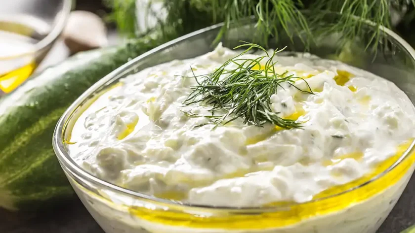 Glasschüssel gefüllt mit Tsatsiki, auf dem Olivenöl schwimmt, garniert mit geschnittenem Dill, im Hintergrund liegen Gurken und Dillzweige