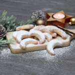 Schön angerichtete, mit Puderzucker bestreute Vanillekipferl in Halbmondform liegen auf einem Brettchen auf dem Tisch. Daneben liegt ein Tattenzweig, kleine goldene Weihnachtsbaumkugeln und ein Teelicht in einem goldenen Stern-Teelichthalter.