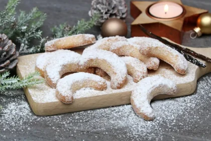 Schön angerichtete, mit Puderzucker bestreute Vanillekipferl in Halbmondform liegen auf einem Brettchen auf dem Tisch. Daneben liegt ein Tattenzweig, kleine goldene Weihnachtsbaumkugeln und ein Teelicht in einem goldenen Stern-Teelichthalter.