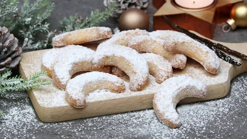 Schön angerichtete, mit Puderzucker bestreute Vanillekipferl in Halbmondform liegen auf einem Brettchen auf dem Tisch. Daneben liegt ein Tattenzweig, kleine goldene Weihnachtsbaumkugeln und ein Teelicht in einem goldenen Stern-Teelichthalter.