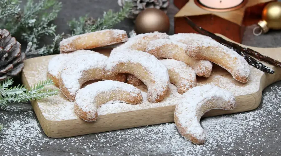 Schön angerichtete, mit Puderzucker bestreute Vanillekipferl in Halbmondform liegen auf einem Brettchen auf dem Tisch. Daneben liegt ein Tattenzweig, kleine goldene Weihnachtsbaumkugeln und ein Teelicht in einem goldenen Stern-Teelichthalter.