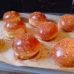 Mehrere frisch gebackene, glänzende, runde Burger-Brötchen auf einem Blech mit Backpapier.