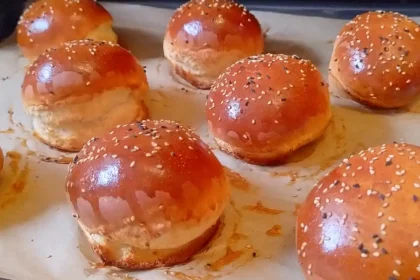 Mehrere frisch gebackene, glänzende, runde Burger-Brötchen auf einem Blech mit Backpapier.
