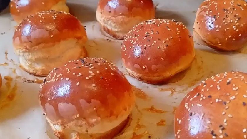 Mehrere frisch gebackene, glänzende, runde Burger-Brötchen auf einem Blech mit Backpapier.