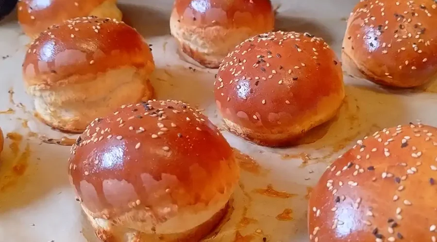 Mehrere frisch gebackene, glänzende, runde Burger-Brötchen auf einem Blech mit Backpapier.