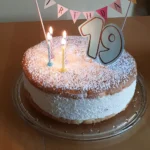 Auf dem Tisch steht auf einer Glas-Tortenplatte eine Torte: Oben und unten ein Bispuitboden, in der Mitte eine hohe Füllung aus Quarkcreme. Die Torte ist bestreut mit Puderzucker, garniert mit vier kleinen, brennenden Kerzen, einer 19 und einer kleinen Wimpelkette auf der „Happy Birthday“ zu lesen ist.