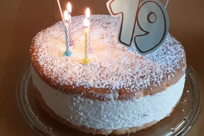 Auf dem Tisch steht auf einer Glas-Tortenplatte eine Torte: Oben und unten ein Bispuitboden, in der Mitte eine hohe Füllung aus Quarkcreme. Die Torte ist bestreut mit Puderzucker, garniert mit vier kleinen, brennenden Kerzen, einer 19 und einer kleinen Wimpelkette auf der „Happy Birthday“ zu lesen ist.