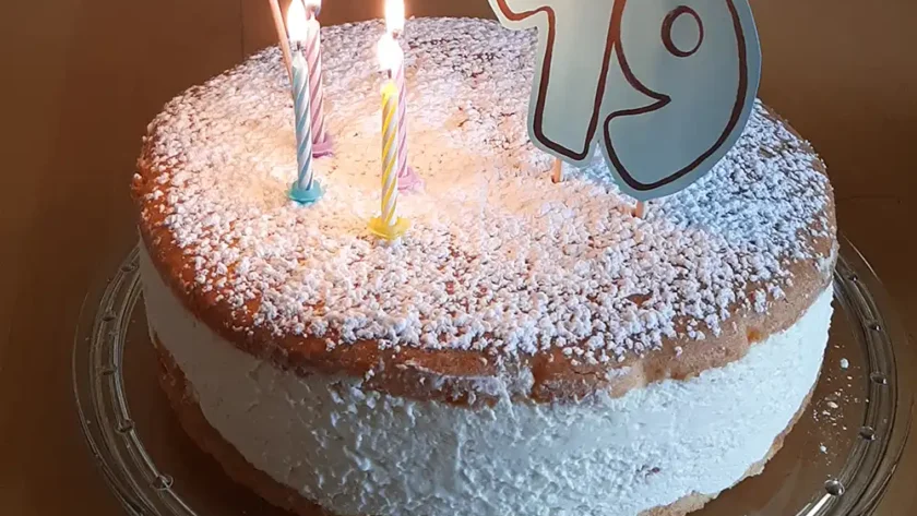 Auf dem Tisch steht auf einer Glas-Tortenplatte eine Torte: Oben und unten ein Bispuitboden, in der Mitte eine hohe Füllung aus Quarkcreme. Die Torte ist bestreut mit Puderzucker, garniert mit vier kleinen, brennenden Kerzen, einer 19 und einer kleinen Wimpelkette auf der „Happy Birthday“ zu lesen ist.