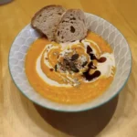 Kürbiscremesuppe in einer Schüssel garniert mit Sahne, Kürbiskernöl und Kürbiskernen, angerichtet mit einer Scheibe Brot