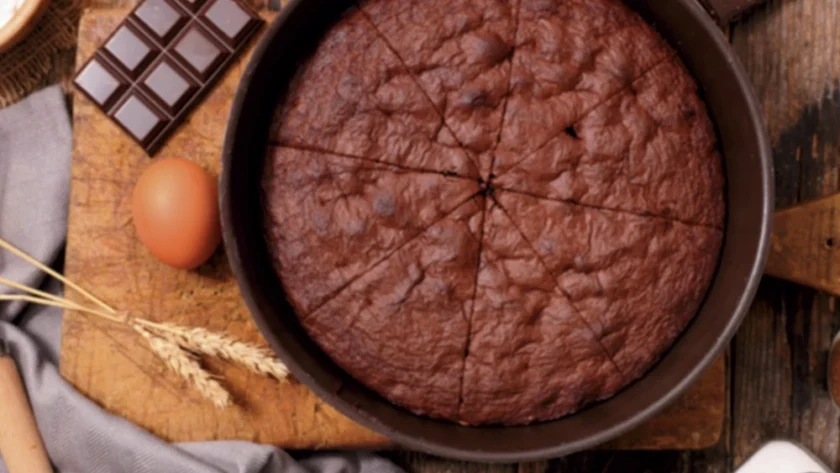 Schokoladen-Rührkuchen in einer runden Backform auf einem Holztisch, daneben liegen Eier und Schokolade