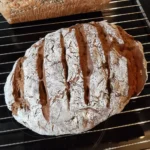 Ein Laib dunkles Brot, mehrfach eingeschnitten und mit Mehl bestreut liegt auf einem Gitter zum Auskühlen.