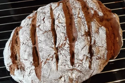 Ein Laib dunkles Brot, mehrfach eingeschnitten und mit Mehl bestreut liegt auf einem Gitter zum Auskühlen.