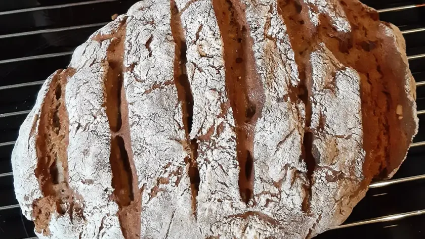 Ein Laib dunkles Brot, mehrfach eingeschnitten und mit Mehl bestreut liegt auf einem Gitter zum Auskühlen.