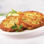 Zwei angebratene Gemüsepuffer auf einem weißen Teller, angerichtet mit Zucchini-Scheiben, garniert mit einem Blatt Petersilie