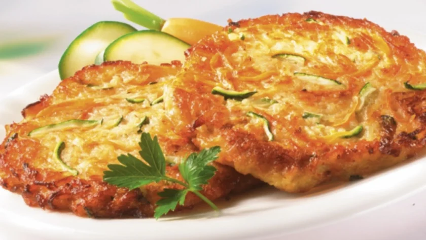 Zwei angebratene Gemüsepuffer auf einem weißen Teller, angerichtet mit Zucchini-Scheiben, garniert mit einem Blatt Petersilie