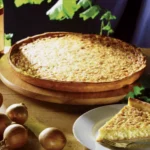 Zwiebelkuchen auf einem Holzbrett im Hintergrund, im >Vordergrund ein Stück Zwiebelkuchen auf einem weißen Teller, daneben liegen ungeschälte Zwiebeln