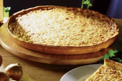 Zwiebelkuchen auf einem Holzbrett im Hintergrund, im >Vordergrund ein Stück Zwiebelkuchen auf einem weißen Teller, daneben liegen ungeschälte Zwiebeln