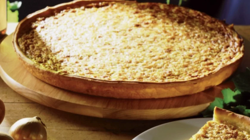 Zwiebelkuchen auf einem Holzbrett im Hintergrund, im >Vordergrund ein Stück Zwiebelkuchen auf einem weißen Teller, daneben liegen ungeschälte Zwiebeln