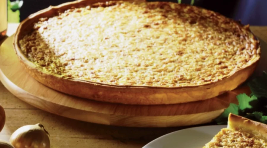Zwiebelkuchen auf einem Holzbrett im Hintergrund, im >Vordergrund ein Stück Zwiebelkuchen auf einem weißen Teller, daneben liegen ungeschälte Zwiebeln