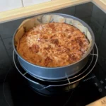 Ein frisch gebackener Kuchen bestreut mit Mandeln steht noch in der runden, mit Backpapier ausgelegten Backform auf einem Kuchengitter auf dem Herd zum Auskühlen.