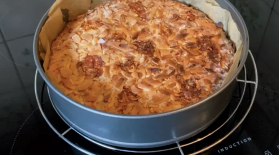 Ein frisch gebackener Kuchen bestreut mit Mandeln steht noch in der runden, mit Backpapier ausgelegten Backform auf einem Kuchengitter auf dem Herd zum Auskühlen.
