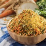 Eine kleine Holzschüssel steht auf dem Tisch und ist bis oben hin gefüllt mit einer Mischung aus getrocknetem, kleingehäckseltem Gemüse. Im Hintergrund liegt eine Sellerieknolle, ein paar Karotten und Petersilie.