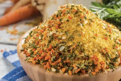 Eine kleine Holzschüssel steht auf dem Tisch und ist bis oben hin gefüllt mit einer Mischung aus getrocknetem, kleingehäckseltem Gemüse. Im Hintergrund liegt eine Sellerieknolle, ein paar Karotten und Petersilie.