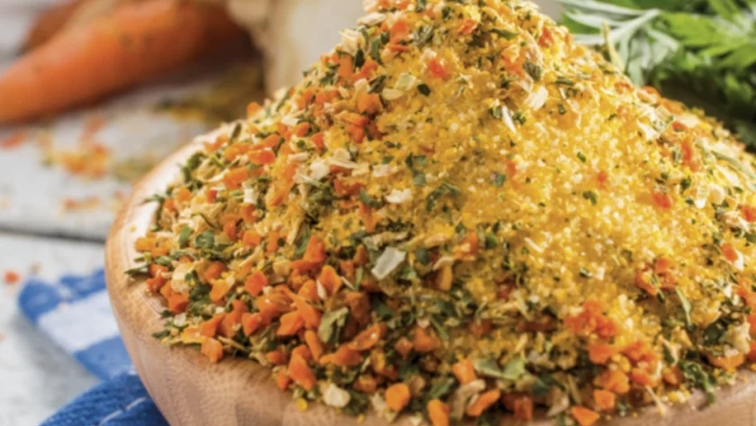 Eine kleine Holzschüssel steht auf dem Tisch und ist bis oben hin gefüllt mit einer Mischung aus getrocknetem, kleingehäckseltem Gemüse. Im Hintergrund liegt eine Sellerieknolle, ein paar Karotten und Petersilie.