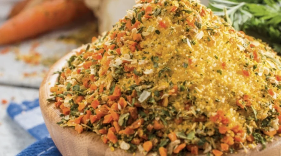 Eine kleine Holzschüssel steht auf dem Tisch und ist bis oben hin gefüllt mit einer Mischung aus getrocknetem, kleingehäckseltem Gemüse. Im Hintergrund liegt eine Sellerieknolle, ein paar Karotten und Petersilie.