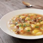 Ein weißer Porzellanteller steht gefüllt mit einem Eintopf aus Kartoffeln, Karotten, Würstchen-Stücken und Petersilie auf einem Holztisch. Daneben liegt ein Löffel.