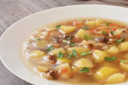 Ein weißer Porzellanteller steht gefüllt mit einem Eintopf aus Kartoffeln, Karotten, Würstchen-Stücken und Petersilie auf einem Holztisch. Daneben liegt ein Löffel.