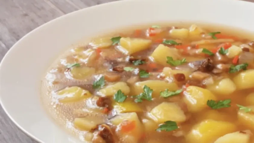 Ein weißer Porzellanteller steht gefüllt mit einem Eintopf aus Kartoffeln, Karotten, Würstchen-Stücken und Petersilie auf einem Holztisch. Daneben liegt ein Löffel.