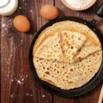 Sicht von oben auf eine Pfanne, in der mehrere Pfannkuchen übereinander gestapelt liegen. Die Pfanne steht auf einem rustikalen dunklen Holztisch. Als Dekoration liegen noch Eier daneben, sowie eine kleine Schüssel Mehl und eine Glaskaraffe mit Milch.