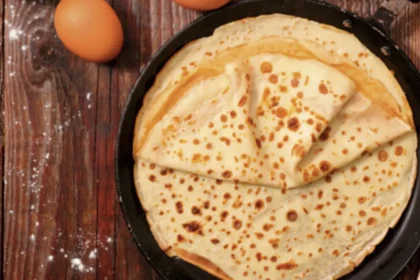 Sicht von oben auf eine Pfanne, in der mehrere Pfannkuchen übereinander gestapelt liegen. Die Pfanne steht auf einem rustikalen dunklen Holztisch. Als Dekoration liegen noch Eier daneben, sowie eine kleine Schüssel Mehl und eine Glaskaraffe mit Milch.
