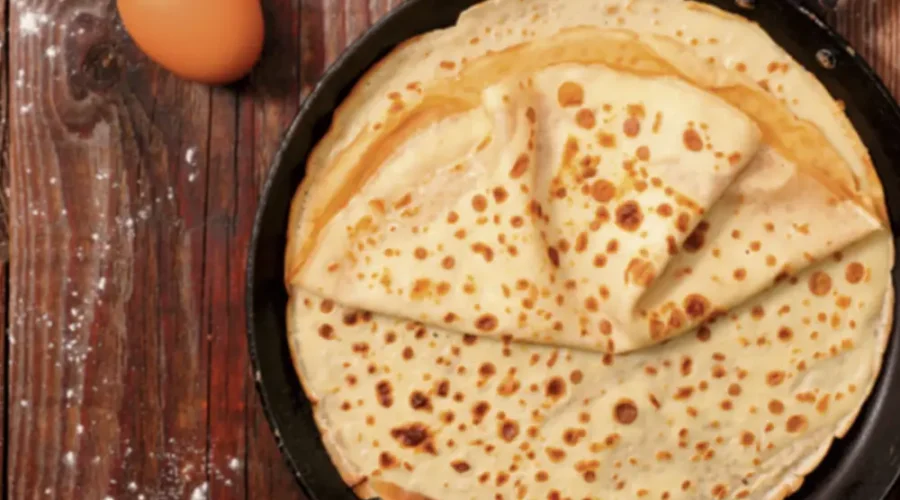 Sicht von oben auf eine Pfanne, in der mehrere Pfannkuchen übereinander gestapelt liegen. Die Pfanne steht auf einem rustikalen dunklen Holztisch. Als Dekoration liegen noch Eier daneben, sowie eine kleine Schüssel Mehl und eine Glaskaraffe mit Milch.
