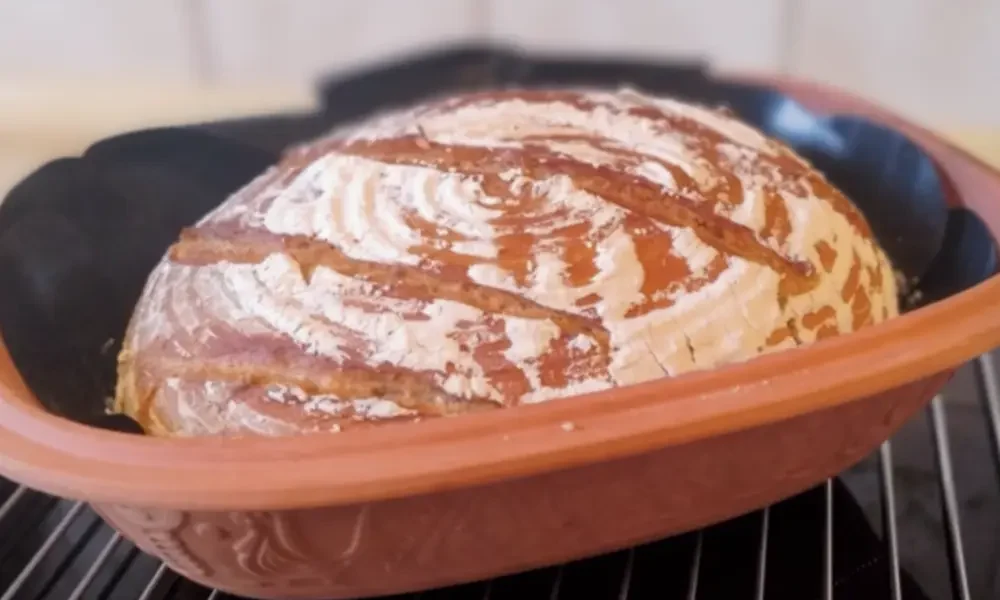 Frisch gebackener Brotlaib mit Mehl auf der Kruste liegt in einem Römertopf zum auskühlen auf einem Gitter.