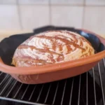 Frisch gebackener Brotlaib mit Mehl auf der Kruste liegt in einem Römertopf zum auskühlen auf einem Gitter.