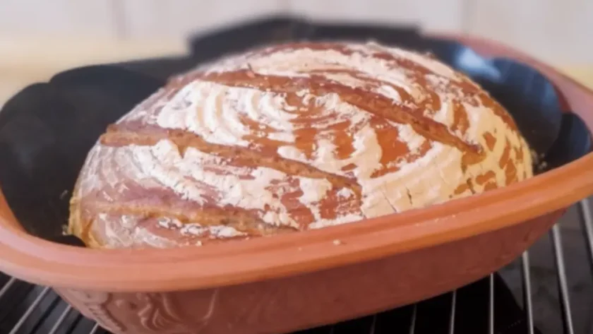 Frisch gebackener Brotlaib mit Mehl auf der Kruste liegt in einem Römertopf zum auskühlen auf einem Gitter.