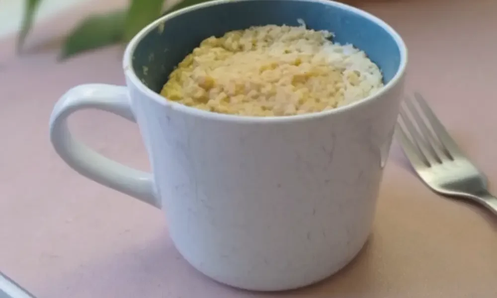 Eine große Tasse steht auf einem Tisch. Die Tasse ist gefüllt mit einem Kuchen, neben der TAsse liegt eine Gabel.