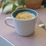 Eine große Tasse steht auf einem Tisch. Die Tasse ist gefüllt mit einem Kuchen, neben der TAsse liegt eine Gabel.