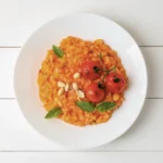 Von oben fotografierter weißer Porzellanteller gefüllt mit tomatigem Risotto, garniert mit gerösteten Rispen-Tomaten.